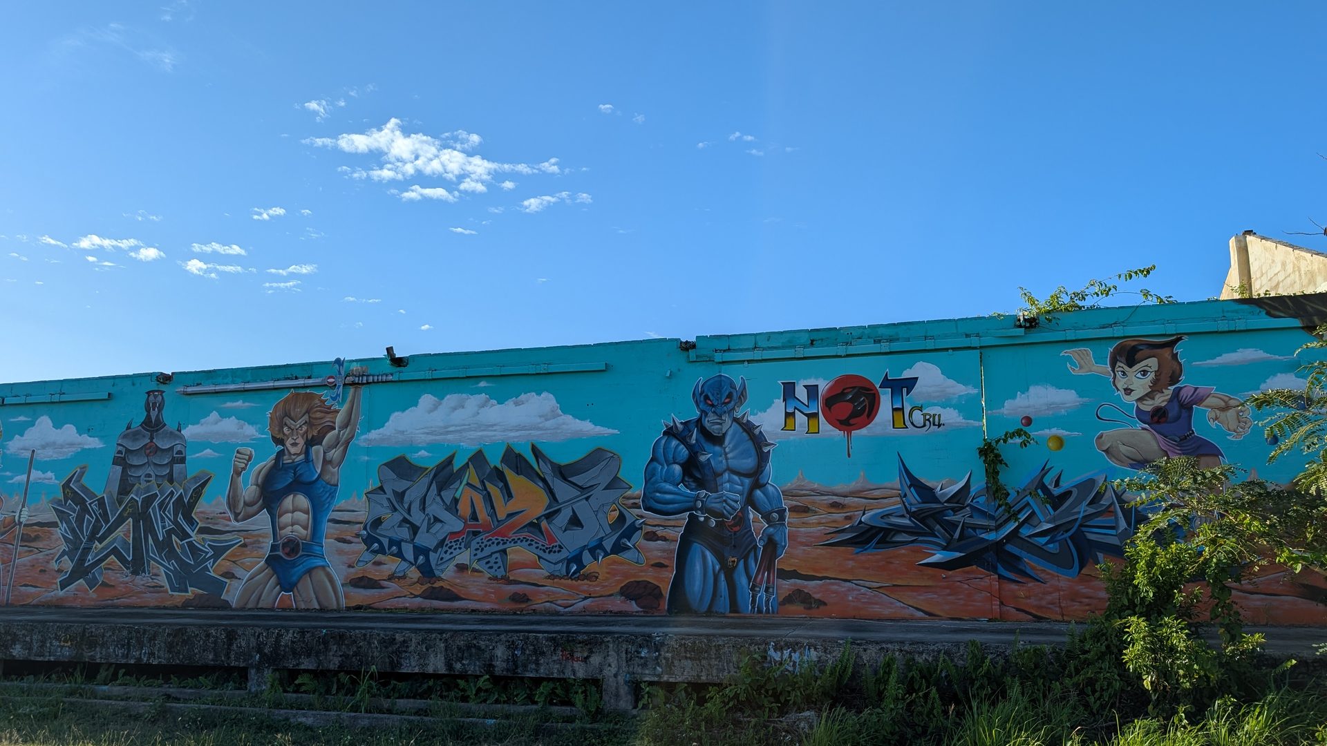 Wide mural of warriors and comic book characters painted on a long teal wall under blue sky